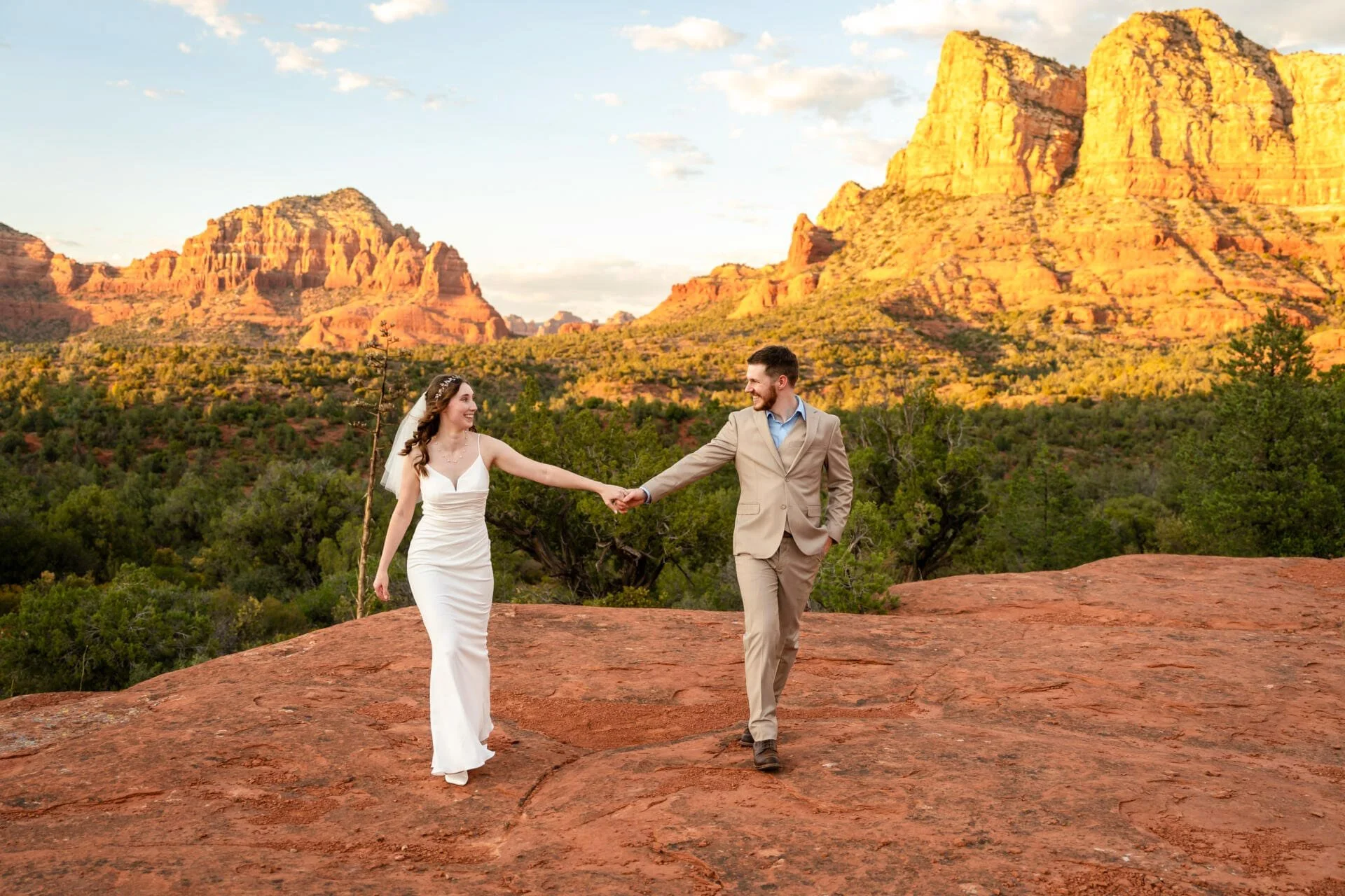 Couple walking hand in hand across red rock below sunlit buttes at golden hour — Sedona, AZ