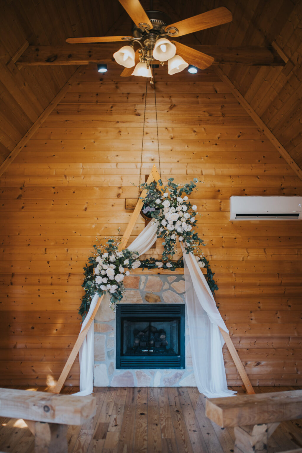 Elopement Wedding A cozy wooden chapel interior perfect for an elopement, featuring a triangular wooden frame adorned with white flowers and greenery above a stone fireplace. Sheer white fabric drapes elegantly over the frame. A wooden ceiling fan with lights hangs above, and wooden benches can be seen in the foreground. Elopements Inc