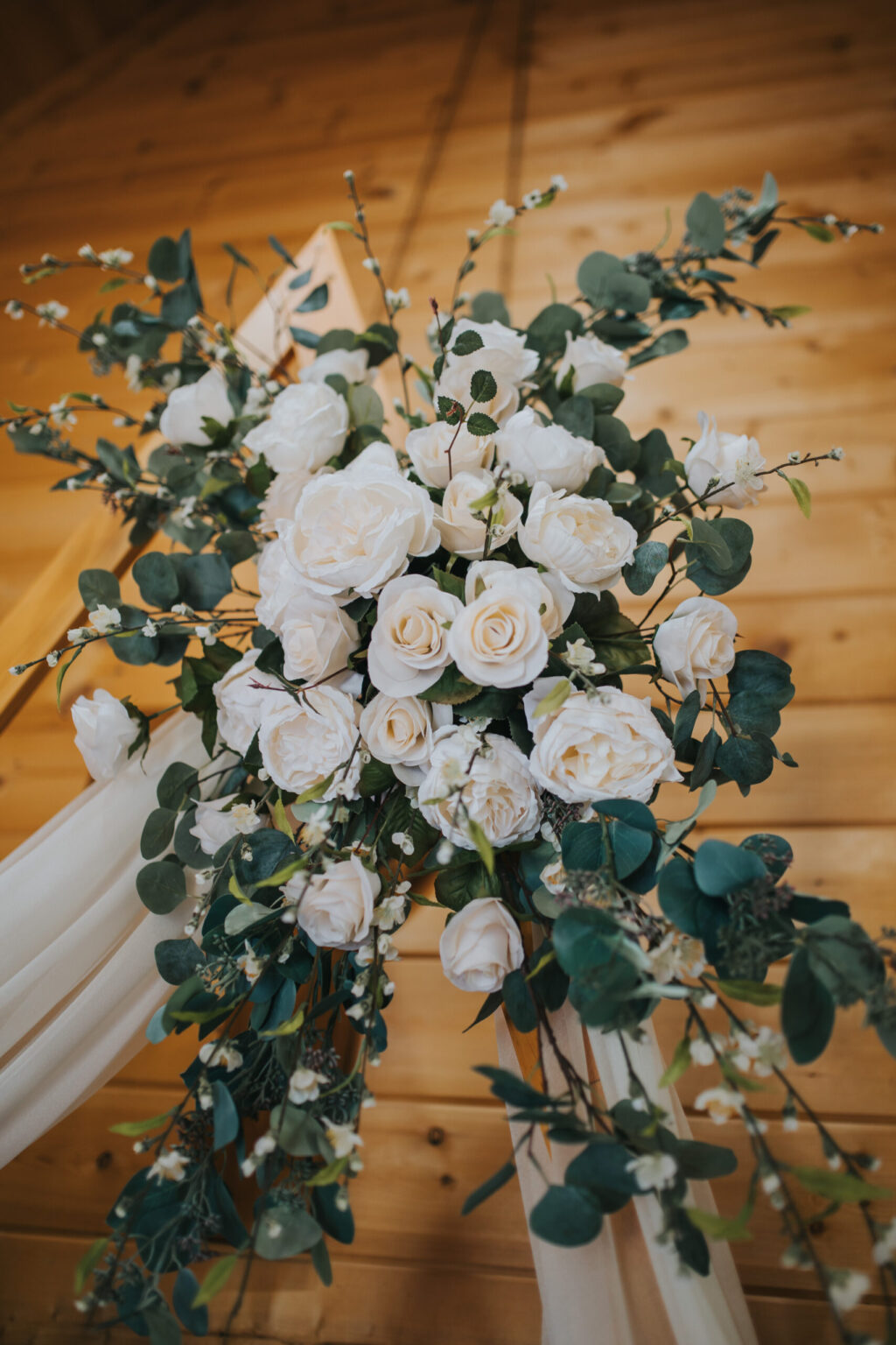 Elopement Wedding A floral arrangement featuring white roses and lush green leaves is displayed against a wooden backdrop, perfect for an intimate elopement. Willow branches add height and texture to the bouquet, while cream-colored fabric drapes gracefully from the arrangement, enhancing the elegant, rustic decor. Elopements Inc