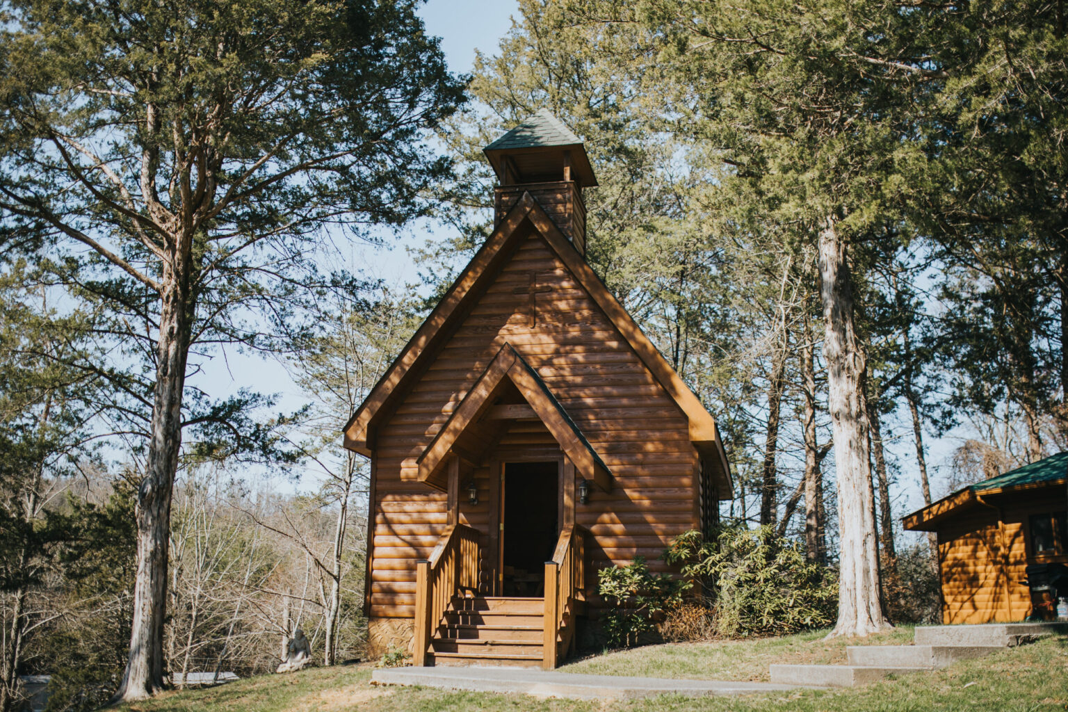 Elopement Wedding A small, rustic wooden cabin surrounded by tall trees under a clear sky—perfect for intimate elopements. The cabin has a triangular roof with a chimney, a front porch with steps, and a wooden door. Sunlight filters through the trees, illuminating the exterior. A smaller structure is partially visible to the right. Elopements Inc