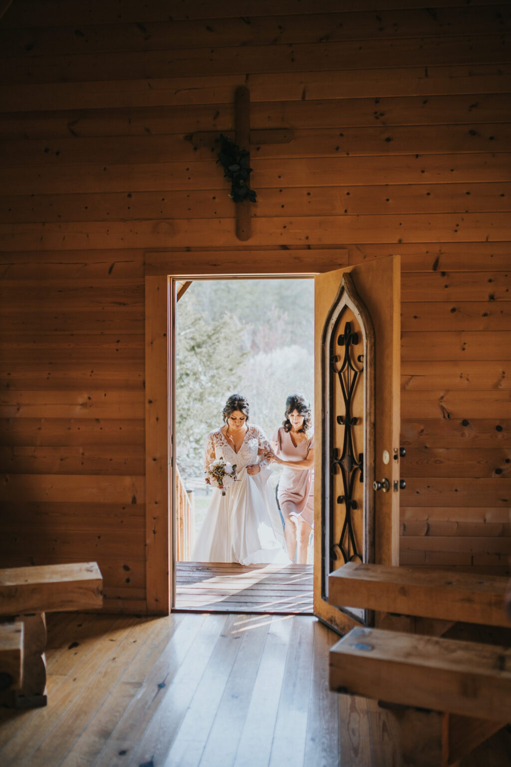 Elopement Wedding Two women are walking through the open door of a wooden chapel. The bride on the left, in a white gown holding a bouquet, and her bridesmaid in light mauve, seem to cherish this intimate elopement. Inside, wooden pews and floors lead to a cross hanging above the entrance. Elopements Inc