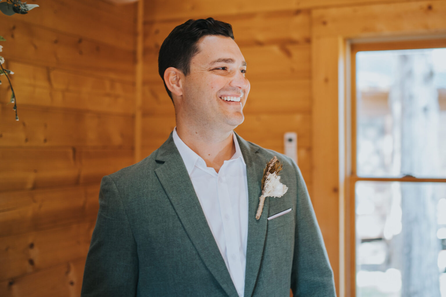 Elopement Wedding A man is smiling broadly while standing indoors in a wooden room, elopement vibes all around. He is wearing a light grey suit, a white dress shirt, and has a feathered boutonniere pinned to his suit jacket. A window with a view of trees is visible in the background. The atmosphere appears warm and inviting. Elopements Inc