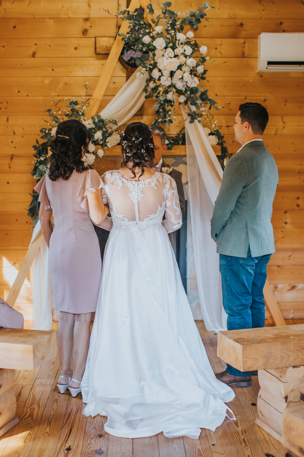 Elopement Wedding A wedding ceremony in a rustic wooden venue. A bride in a white gown with lace details and her hair in an updo stands facing the officiant. A woman in a pink dress and a man in a green blazer and jeans stand beside her. A floral arch with white flowers and greenery frames this intimate elopement perfectly. Elopements Inc