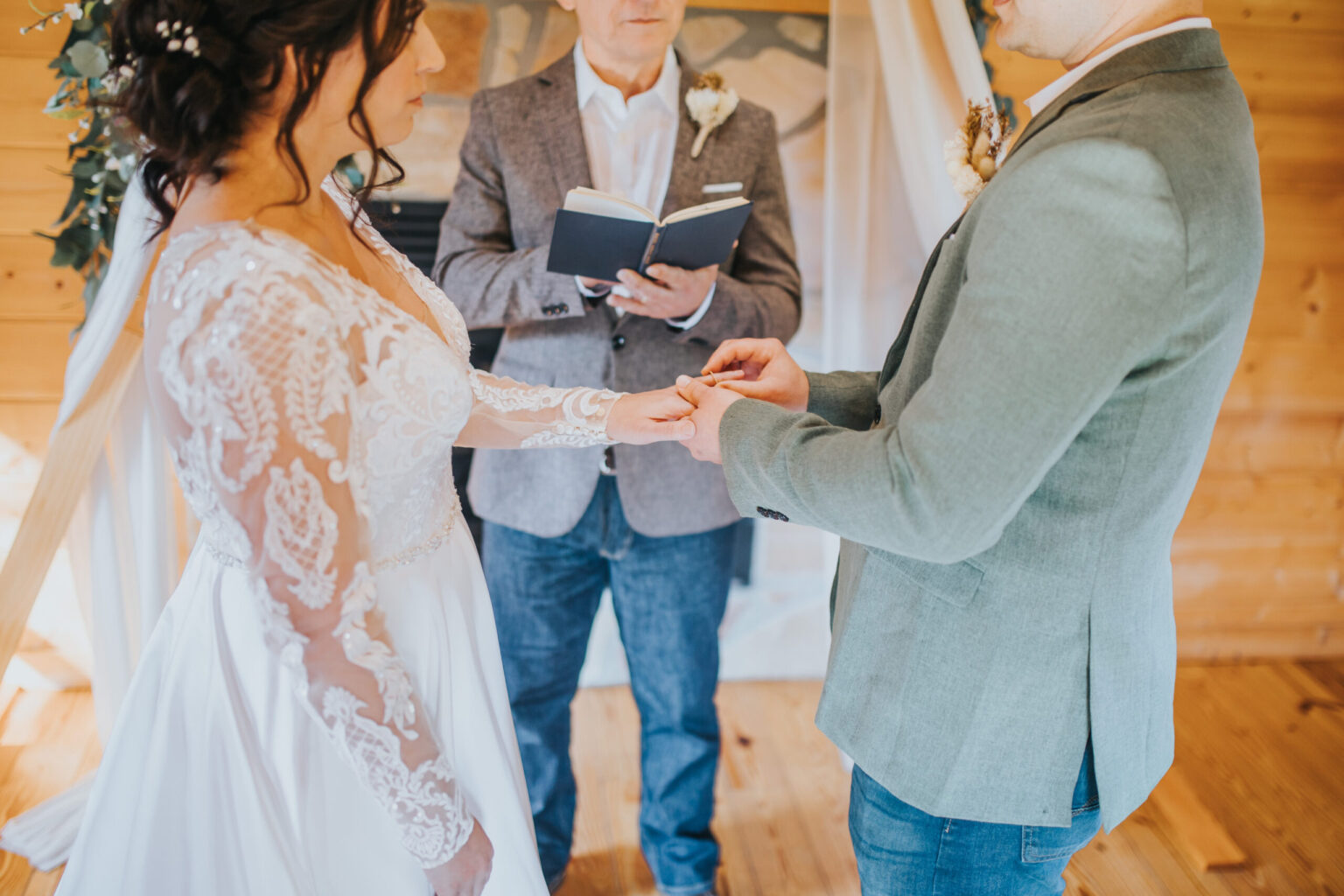 Elopement Wedding A bride and groom, perhaps choosing to elope, are exchanging rings in a wooden interior setting during an intimate wedding ceremony. The bride wears a white lace-trimmed dress, and the groom is dressed in a grey suit jacket. An officiant stands between them, holding an open book with a white bouquet pinned to his jacket. Elopements Inc