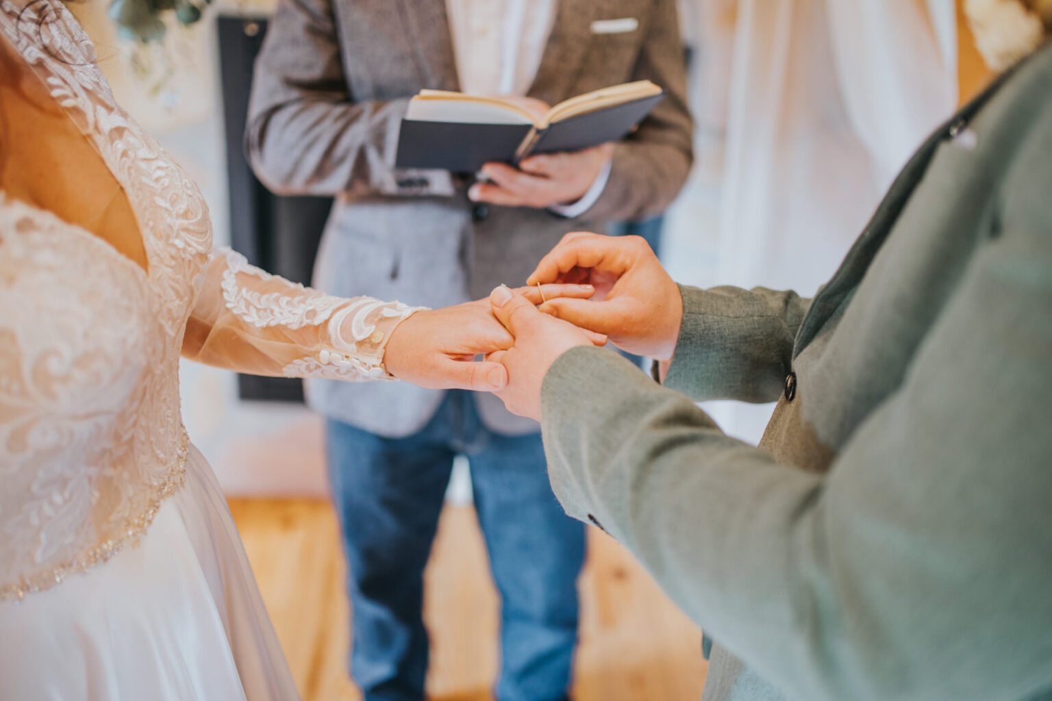 Elopement Wedding A close-up of a wedding ceremony featuring a bride and groom exchanging rings. The bride wears a long-sleeve lace wedding dress, while the groom dons a light grey suit. In the background, an officiant holds an open book, overseeing their elopement. The setting appears intimate and warmly lit. Elopements Inc