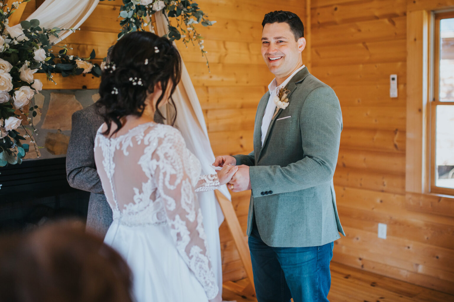 Elopement Wedding A couple elopes in a wooden cabin adorned with flowers and white drapery. The groom, wearing a light gray blazer, smiles as he holds the bride's hand. The bride, in a white lace gown with long sleeves, stands facing him with her back partially visible. Elopements Inc