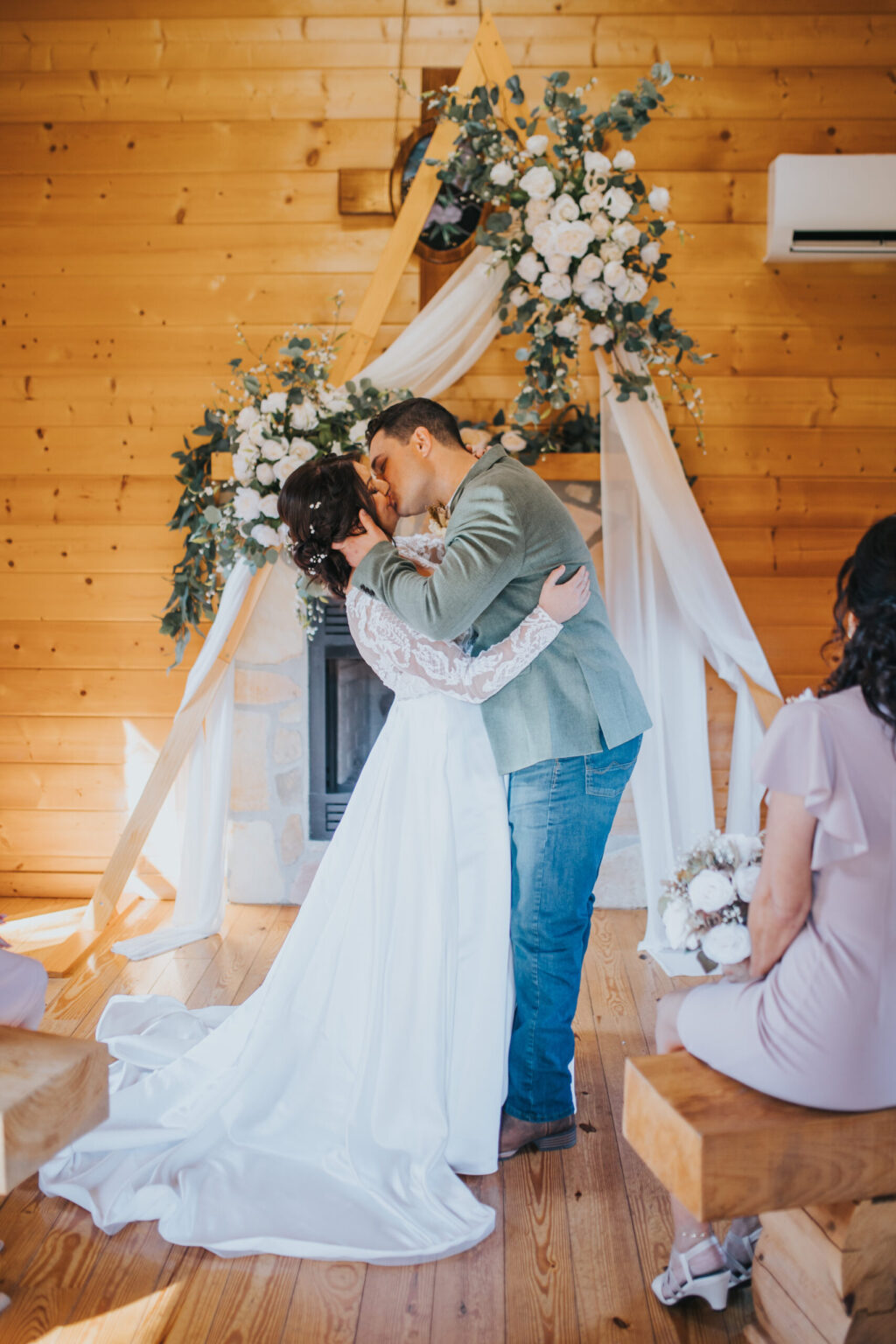 Elopement Wedding A bride and groom share a kiss during their intimate elopement ceremony in a wooden chapel adorned with white floral arrangements and draped fabric. The bride wears a long white dress with lace sleeves, while the groom is in a light gray suit and jeans. Guests are seated on wooden benches, witnessing the moment. Elopements Inc