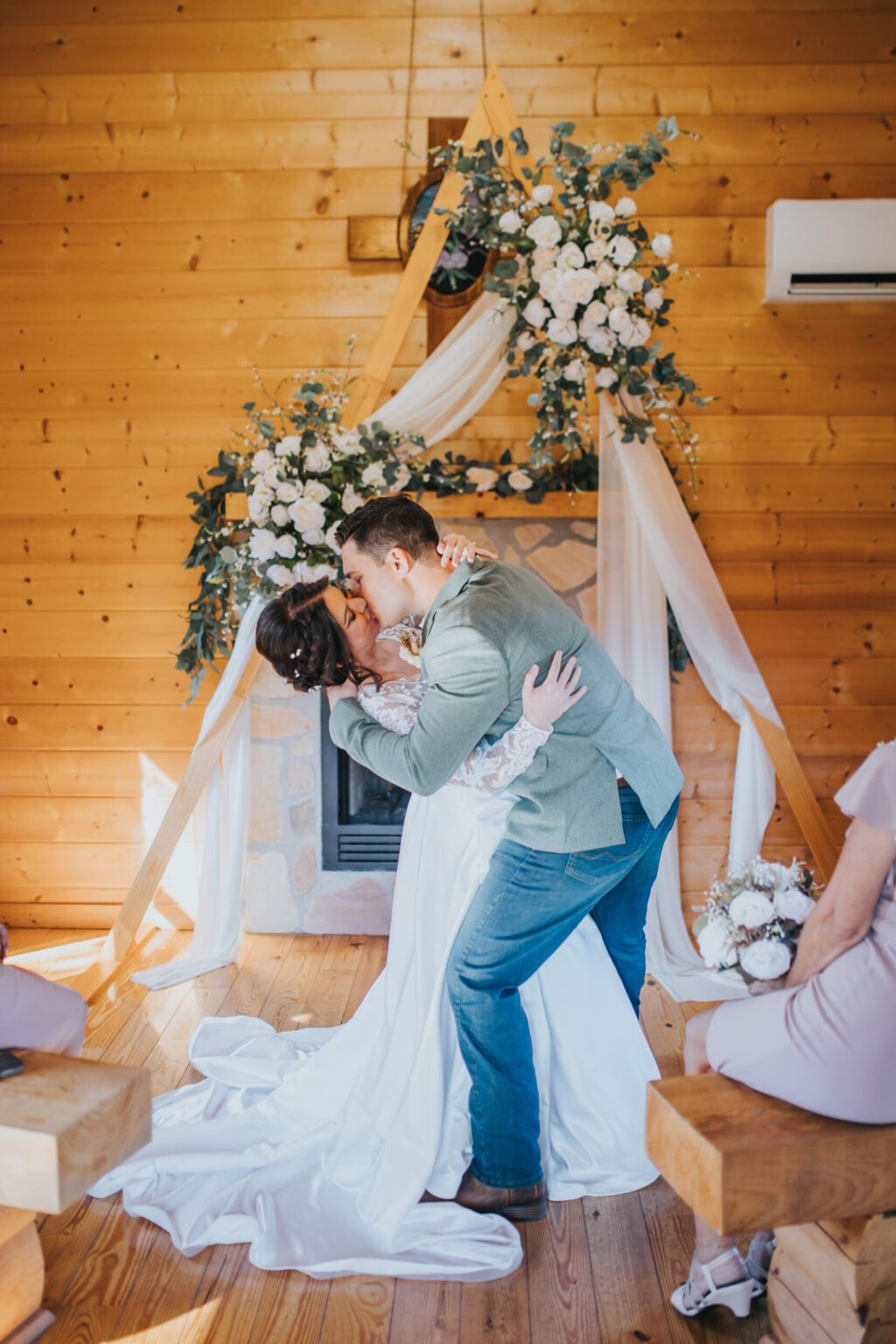 Elopement Wedding A couple shares a passionate kiss at their wedding in a rustic wooden hall adorned with a floral arch and draped white fabric. The bride, in a white gown with a train, and the groom, in a light grey suit, create an intimate moment reminiscent of elopements. Bridesmaids in lavender dresses look on from wooden benches. Elopements Inc