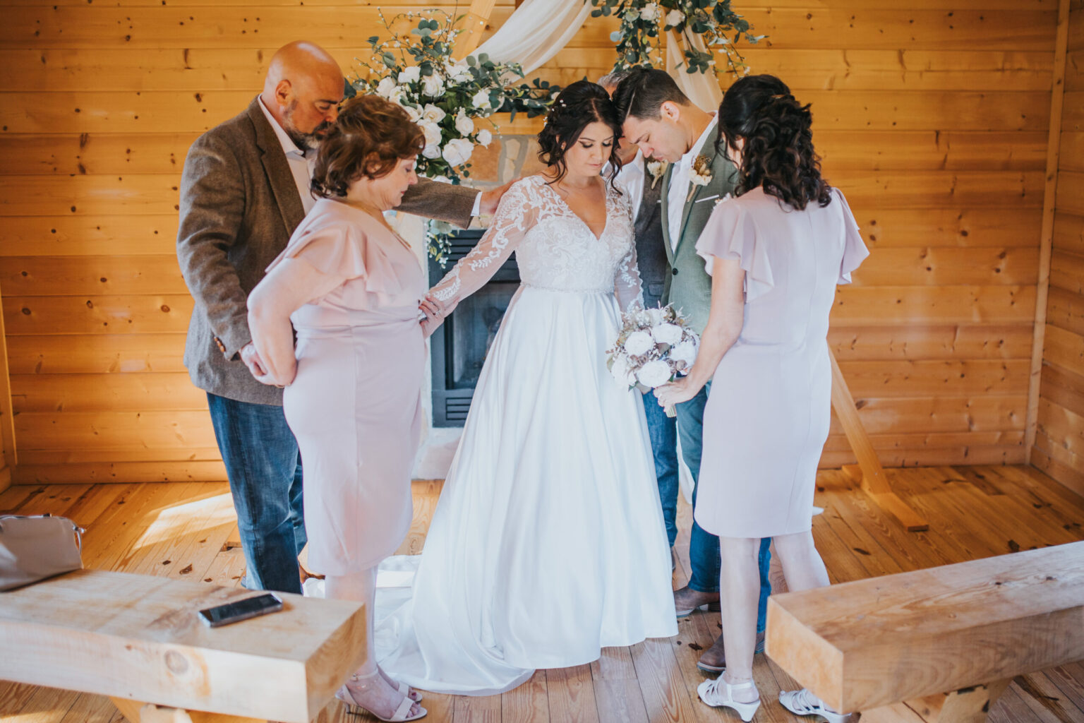 Elopement Wedding A bride in a white wedding gown and groom in a suit stand in a wooden room, surrounded by two women and a man. They join hands and bow their heads in a solemn moment, celebrating their elopement. The room is decorated with white flowers and greenery. Elopements Inc