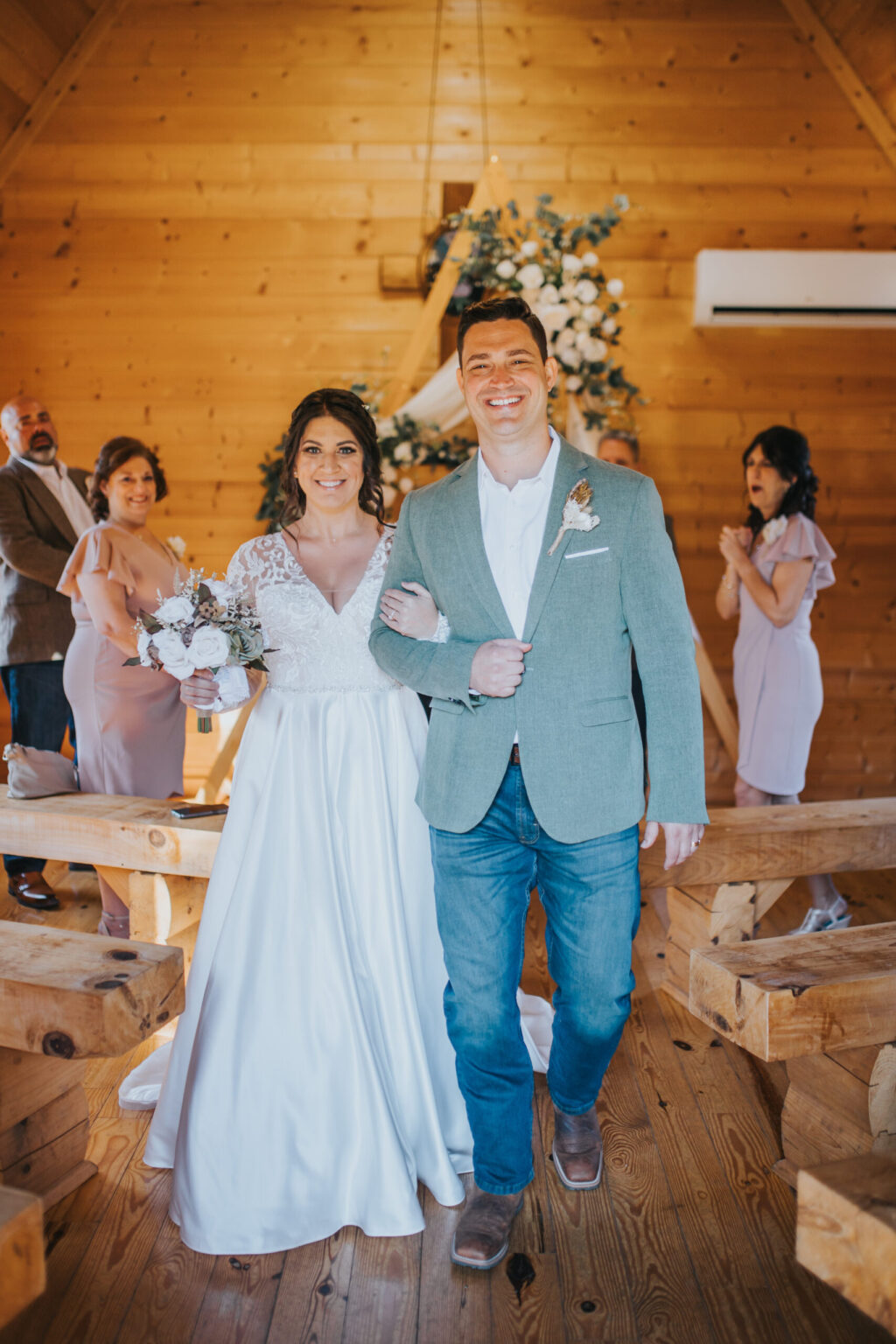 Elopement Wedding A bride in a white gown holds a bouquet and walks arm-in-arm with a groom in a light green jacket and jeans. They are smiling and walking down an aisle in a wooden chapel. Guests, standing and cheering, are visible in the blurred background, celebrating their intimate elopement. Elopements Inc