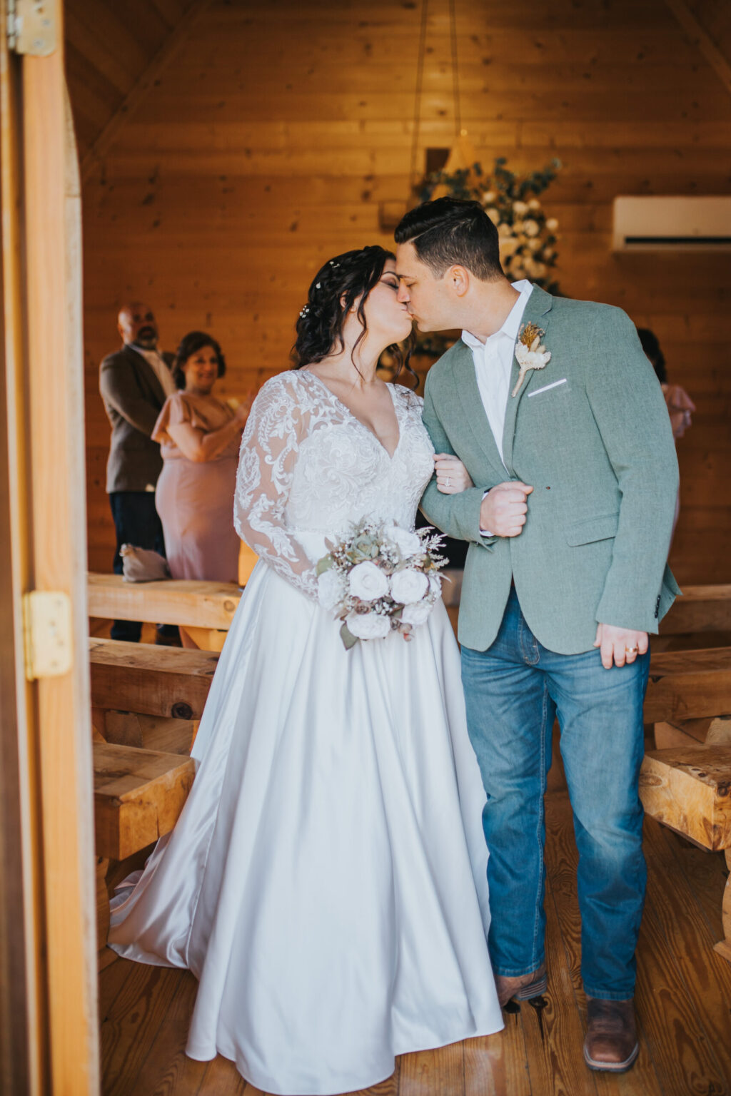 Elopement Wedding A couple shares a kiss in a wooden chapel, having chosen to elope. The bride wears a white lace wedding dress and holds a bouquet of white flowers, while the groom wears a light green blazer, white shirt, and jeans. In the background, guests, including a man and two women, smile and watch the couple. Elopements Inc