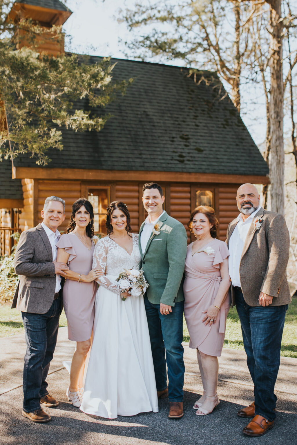 Elopement Wedding A bride in a white gown and a groom in a green jacket, glowing with the joy of their elopement, stand together, smiling, in front of a rustic wooden building. Flanked by three men and two women in semi-formal attire, the group poses on a sunlit, tree-lined path, creating a warm, celebratory atmosphere. Elopements Inc