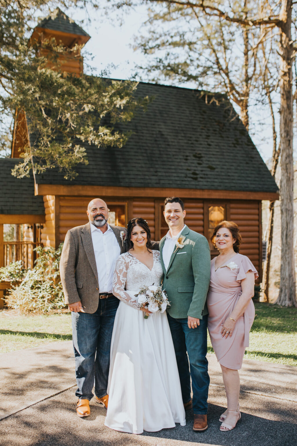 Elopement Wedding A bride and groom, having chosen to elope, stand together, smiling outside a rustic wooden chapel. The bride, in a white lace dress, holds a bouquet. The groom is in a light green jacket and jeans. An older man in a brown jacket and jeans and a woman in a light pink dress stand beside them. Trees surround them. Elopements Inc