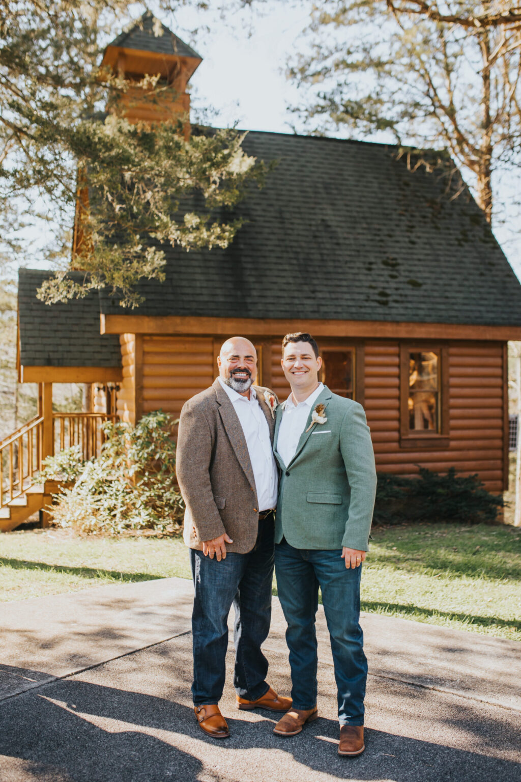 Elopement Wedding Two men, standing closely together and smiling, pose for a photo in front of a rustic wooden cabin. Both wear jeans and dress shirts with sports jackets, one in gray and the other in green. The setting is sunny with trees and a grassy area, creating a warm outdoor atmosphere perfect for an elopement. Elopements Inc