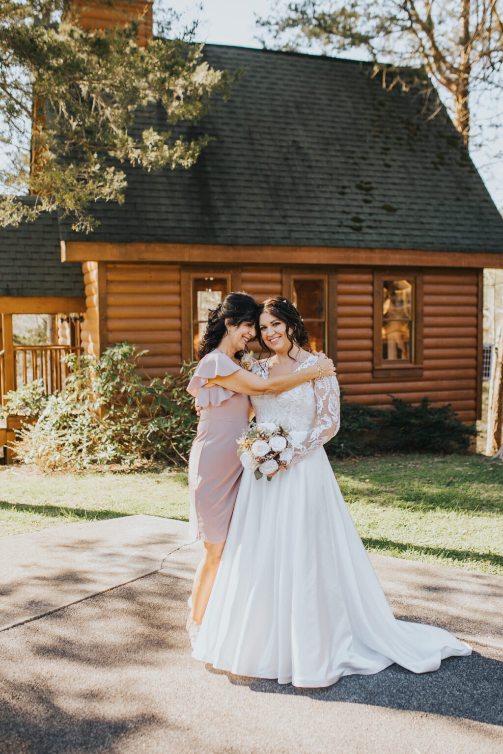 Elopement Wedding Two women, one in a light purple dress and the other in a white wedding gown, smile and embrace in front of a rustic log cabin surrounded by greenery. The bride holds a bouquet of flowers. Their spontaneous elopement is illuminated by sunlight, casting soft shadows and creating a warm atmosphere. Elopements Inc