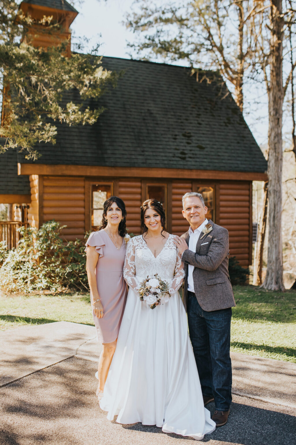 Elopement Wedding A bride in a white wedding dress, holding a bouquet, stands between two older adults. The woman on the left wears a light pink dress, and the man on the right is in a gray blazer and white shirt. They are outdoors, eloping in front of a wooden cabin with trees and greenery in the background. Elopements Inc