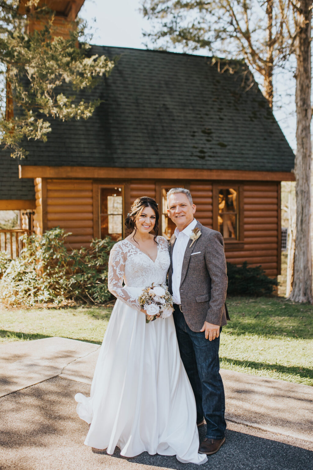 Elopement Wedding A bride in a white dress with lace sleeves and a groom in a grey blazer and jeans stand close together on a driveway, smiling and holding a bouquet. Behind them is a log cabin with a green roof and several trees, suggesting an intimate elopement in a rustic, outdoor setting. Elopements Inc