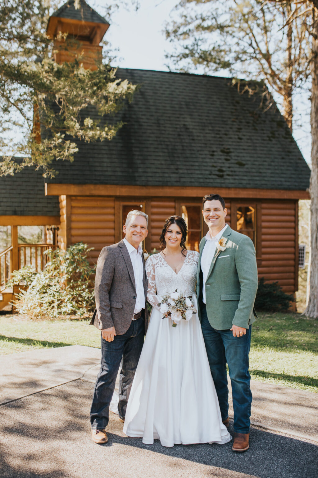 Elopement Wedding Three people stand in front of a rustic wooden chapel, perfect for an intimate elopement. A woman in a white wedding gown with lace sleeves holds a flower bouquet, flanked by two men in blazers. One man wears a gray blazer, and the other wears a green one with a boutonniere. Trees and greenery surround the chapel. Elopements Inc