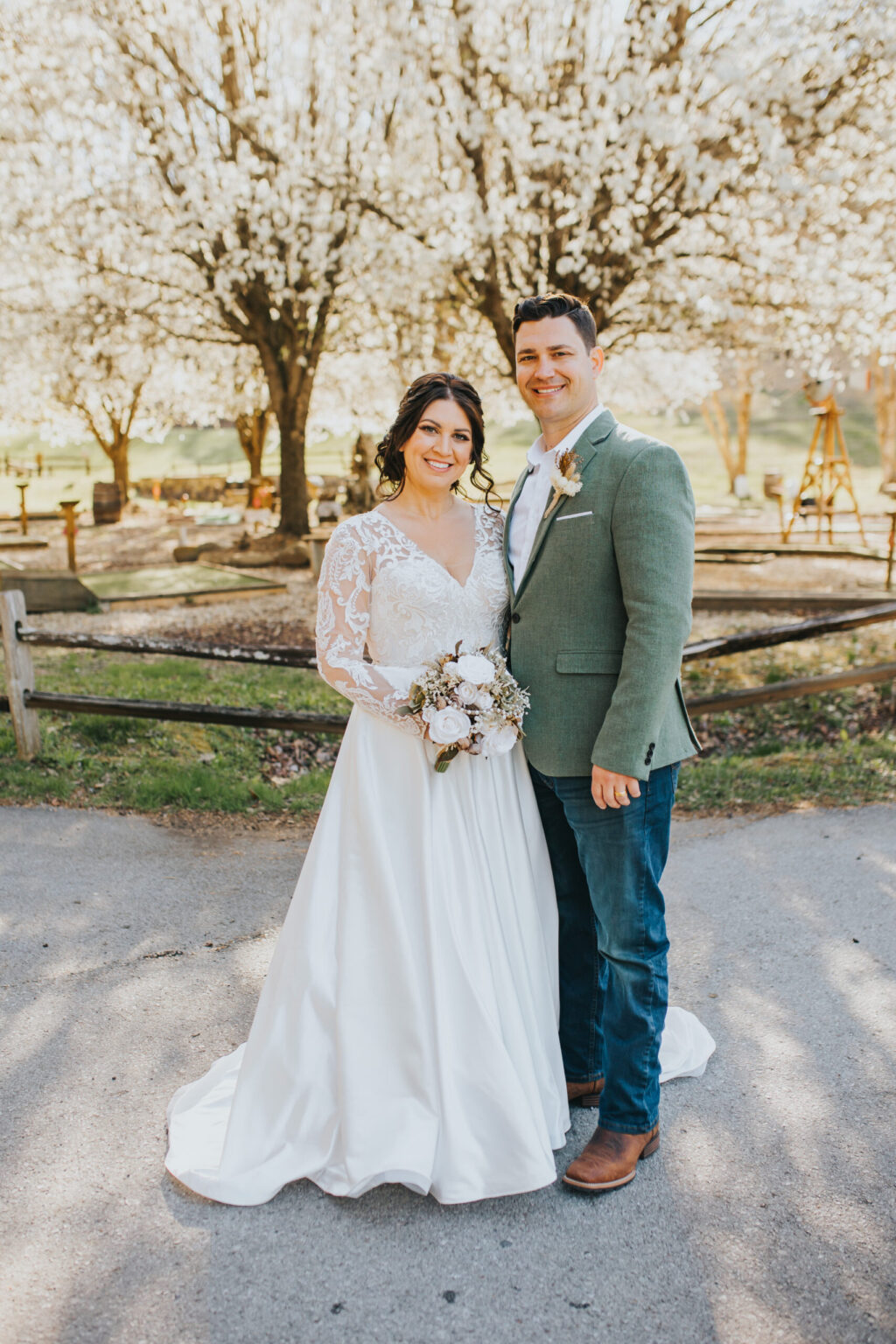 Elopement Wedding A smiling bride and groom who have just decided to elope stand outdoors in front of a blossoming tree. The bride wears a white lace gown and holds a bouquet, while the groom is in a green jacket and jeans, with one arm around her. They are standing on a paved path near a rustic wooden fence in a sunlit, rural setting. Elopements Inc