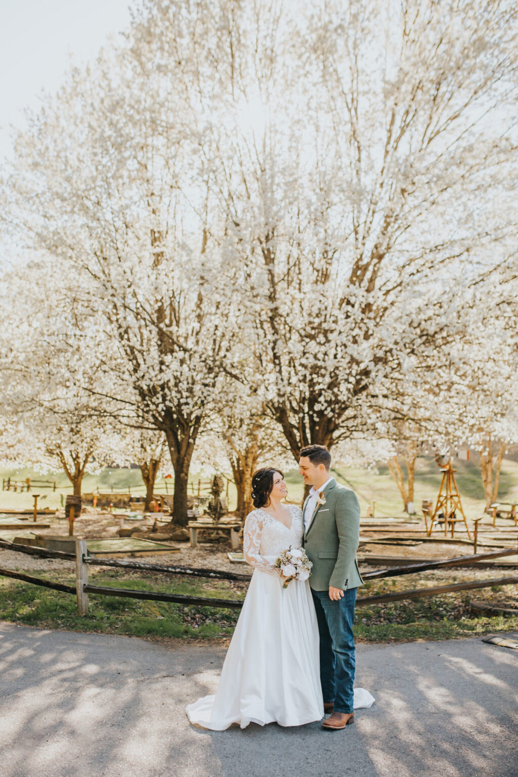 Elopement Wedding A bride and groom stand close together, gazing into each other's eyes, embodying the essence of a romantic elopement. The bride wears a long white gown, and the groom is in a light gray suit. Blooming trees with white flowers provide a lush backdrop, and a wooden fence runs behind them on a sunny day. Elopements Inc