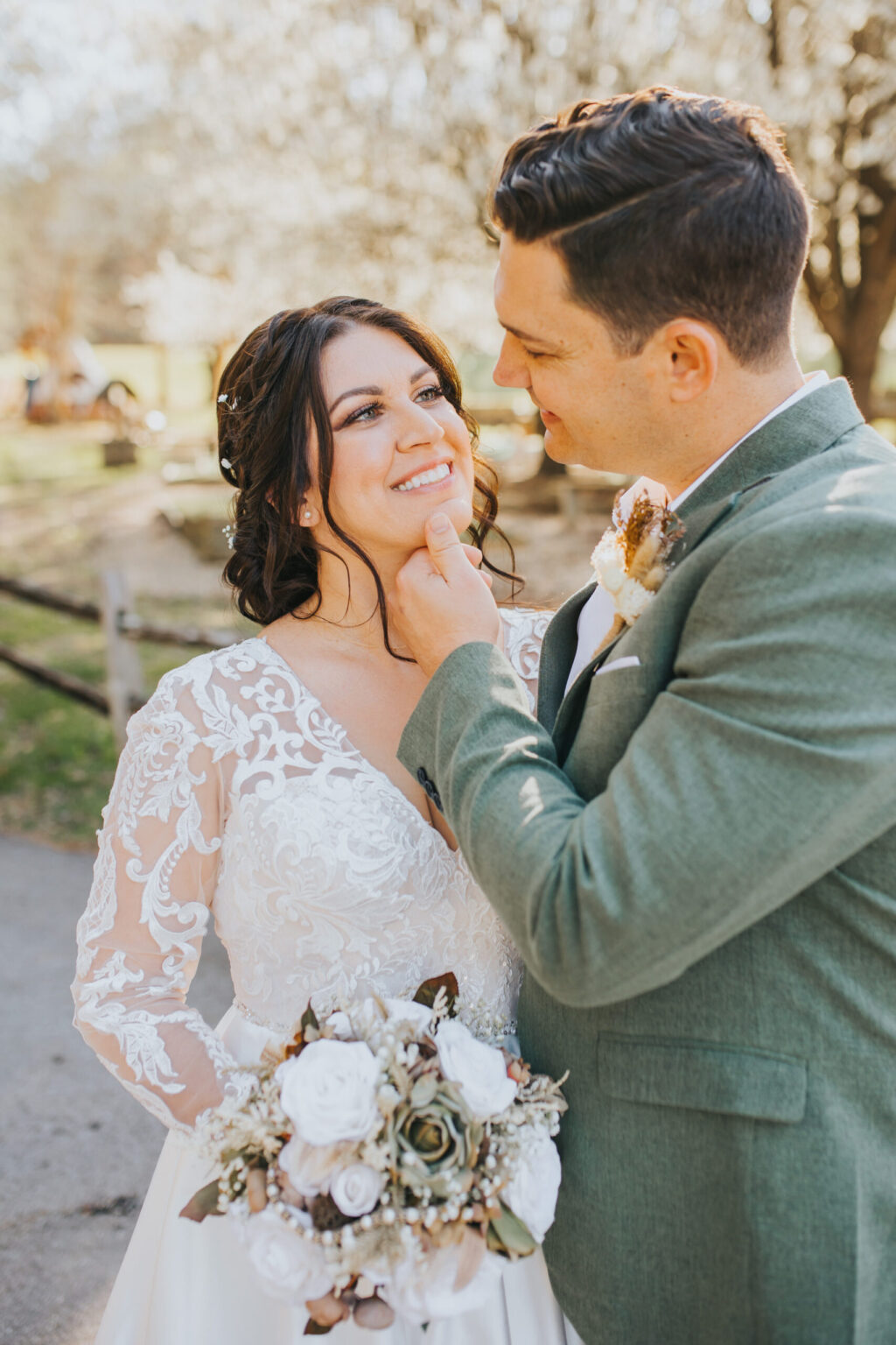 Elopement Wedding A bride and groom share a moment outdoors on their wedding day, almost as if they were eloping. The bride, in a white lace gown, smiles as the groom, in a green suit, gently touches her chin. She holds a bouquet of white flowers and greenery against the backdrop of blurred trees and a wooden fence. Elopements Inc