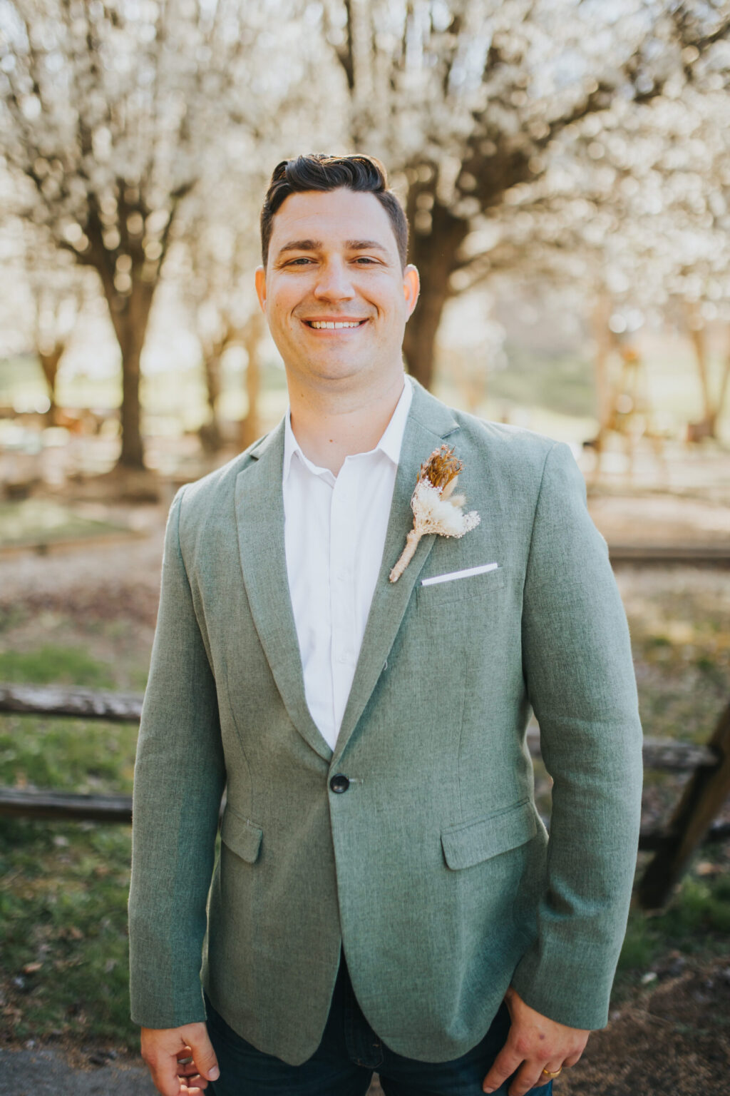 Elopement Wedding A man with short dark hair is smiling and standing outdoors in front of blooming trees. He is wearing a light green blazer over a white shirt, with a brown and white boutonniere pinned to his blazer. The serene setting hints at an intimate elopement, with a wooden fence and green grass in the background. Elopements Inc