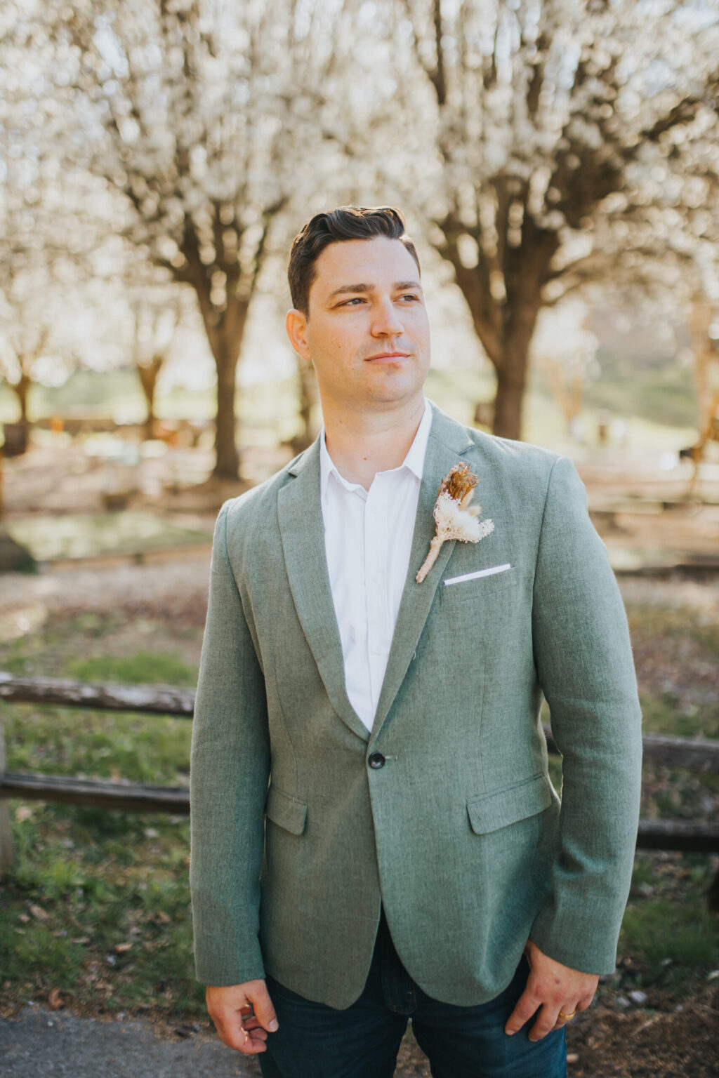 Elopement Wedding A man stands outdoors in a park with blossoming trees in the background, as if ready for an intimate elopement. He is wearing a light green blazer, white shirt, and blue jeans. A small boutonniere with cream and brown flowers is pinned to his left lapel. He gazes slightly to the right with a calm expression. Elopements Inc