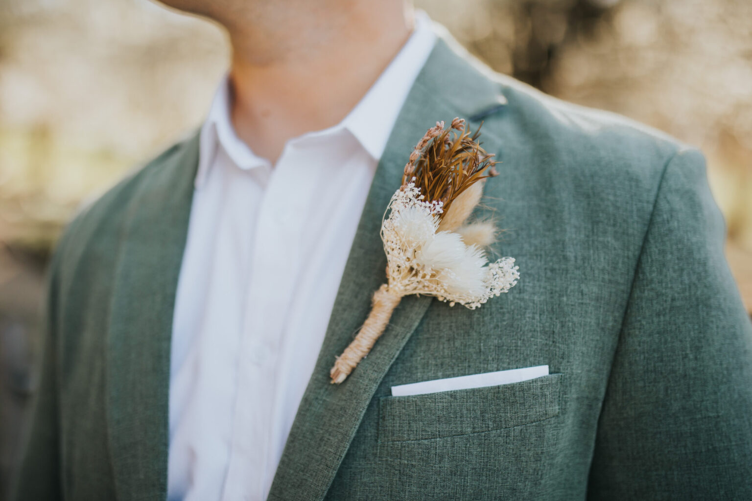 Elopement Wedding Close-up of a person wearing a light green jacket with a white dress shirt underneath, focusing on a rustic boutonniere pinned to the lapel. Perfect for an intimate elopement, the boutonniere features dried flowers and greenery, including white and brown elements, with a small white pocket square visible. Elopements Inc