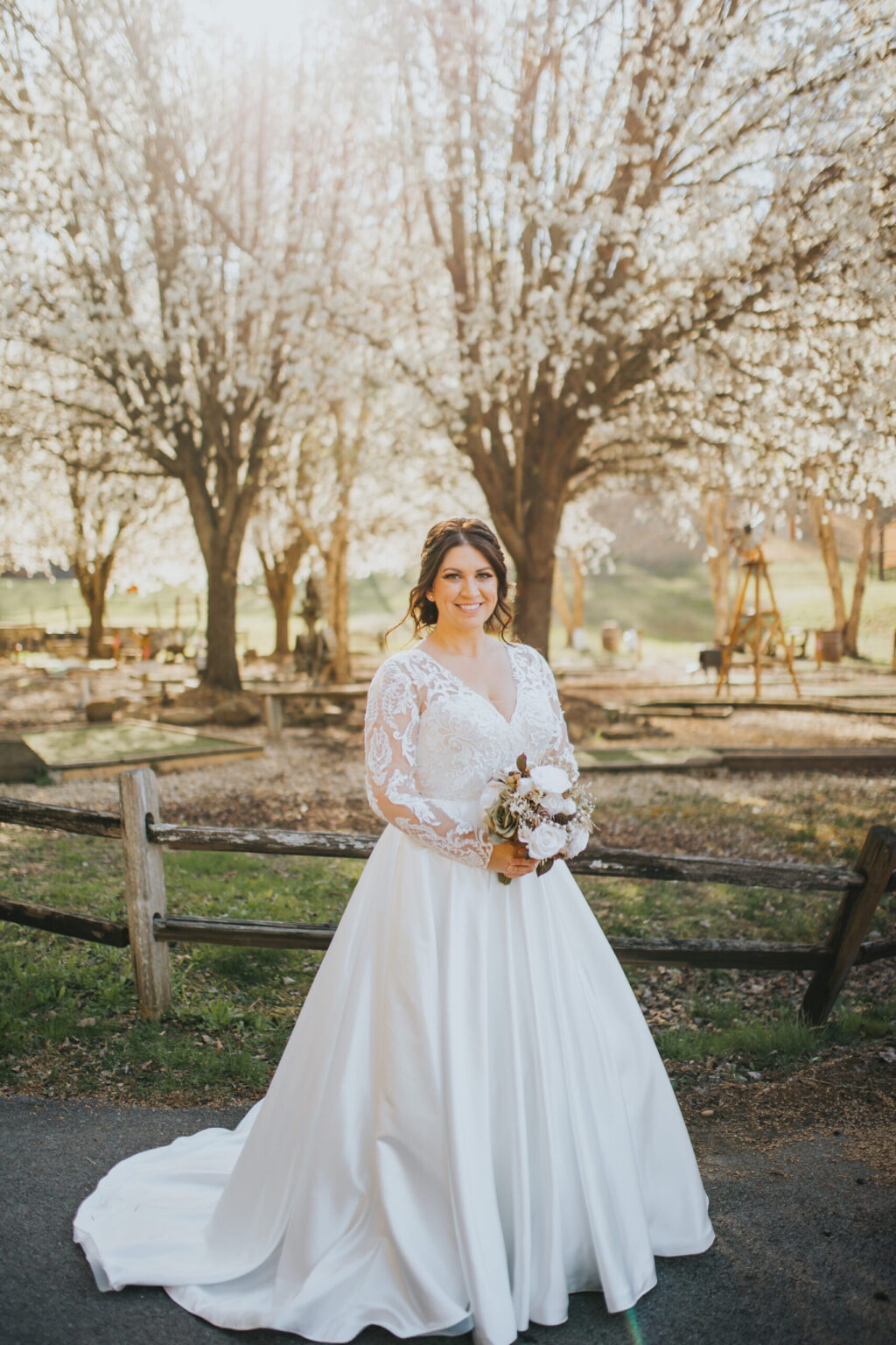 Elopement Wedding A bride wearing a long-sleeved, lace wedding gown stands outdoors holding a bouquet of flowers. Behind her, trees in full bloom create a picturesque background with white blossoms. She is smiling, and wooden fencing and a pathway are visible in the foreground, enhancing the rustic elopement setting. Elopements Inc