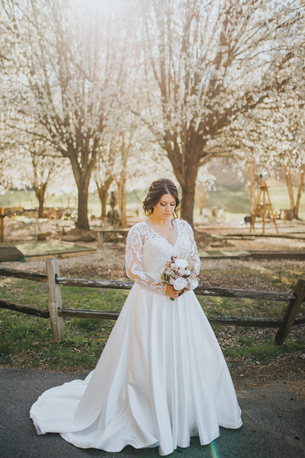 Elopement Wedding A bride in a white wedding dress stands outdoors, holding a bouquet of flowers. The dress features long lace sleeves. Behind her are blooming trees and a wooden fence, with sunlight filtering through the branches, creating a serene and picturesque backdrop perfect for an elopement. The bride gazes down at the bouquet. Elopements Inc