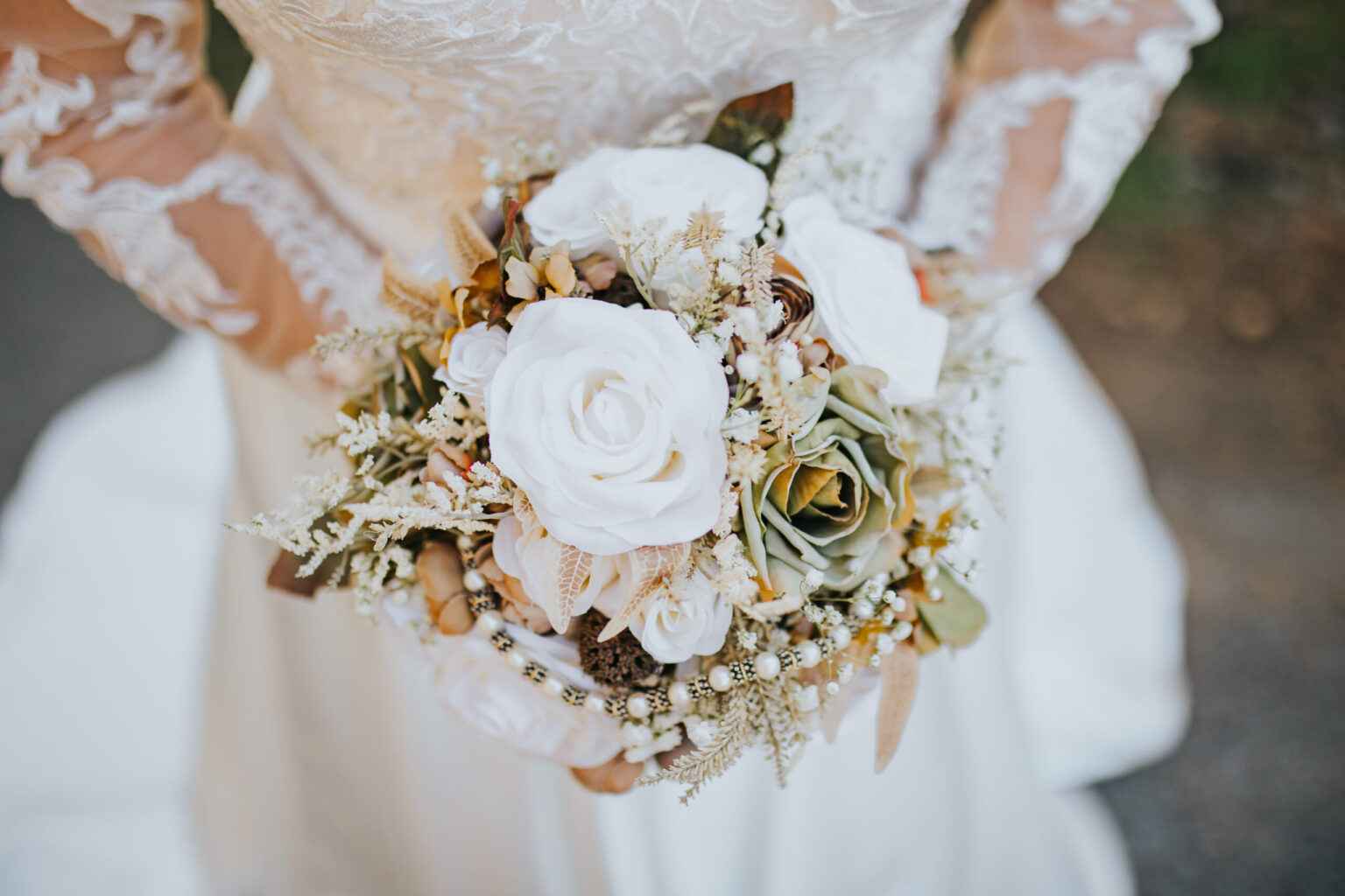 Elopement Wedding Close-up of a bride in a white lace wedding dress holding a bouquet. The bouquet features a variety of white roses, ivory flowers, and greenery, with delicate accents perfect for intimate elopements. The bride's dress has intricate lace sleeves and the background is softly blurred. Elopements Inc