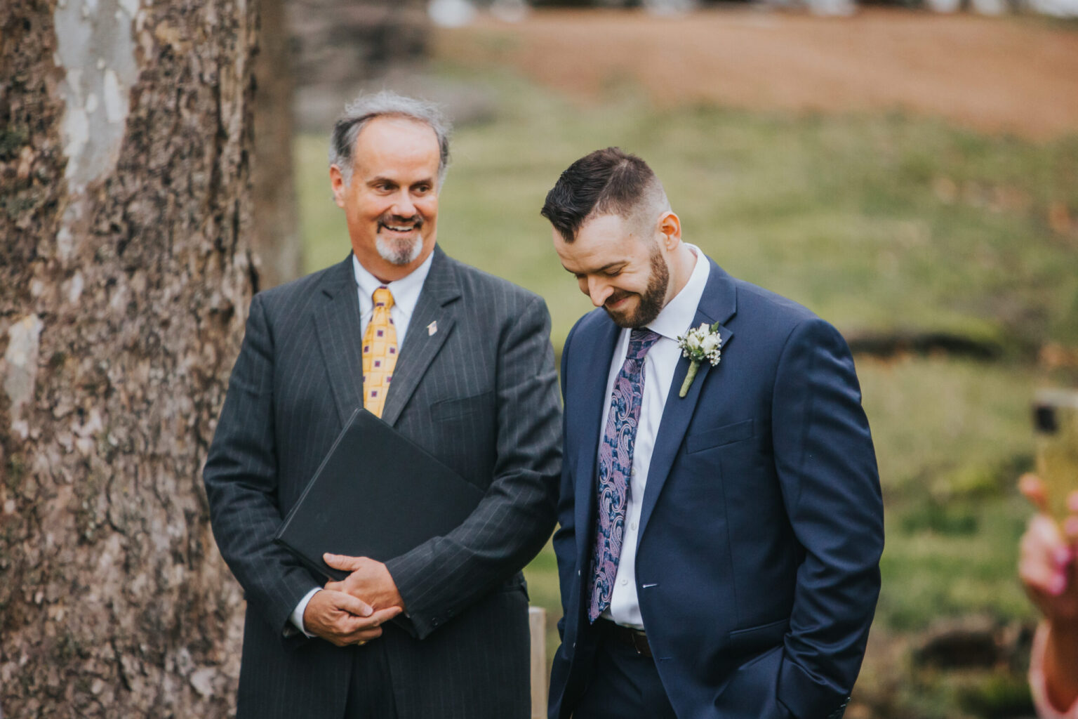 Elopement Wedding A man in a suit and tie holding a folder stands beside another man who is smiling, looking down, and wearing a navy blue suit with a floral tie and boutonniere. They are outdoors, with a tree trunk and blurred greenery in the background, suggesting an elopement ceremony. Elopements Inc