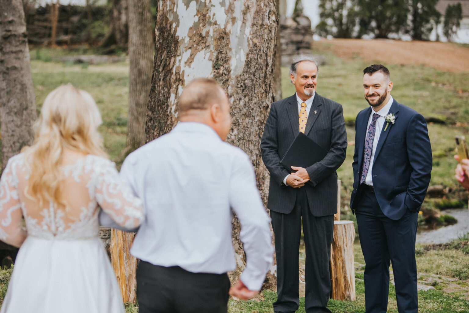 Elopement Wedding A groom in a navy suit with a boutonniere stands next to an officiant in a suit holding a black folder. They are smiling and looking at a bride in a white lace dress and a man walking towards them with their backs to the camera, capturing the intimate moment of their outdoor elopement amidst grass and trees. Elopements Inc