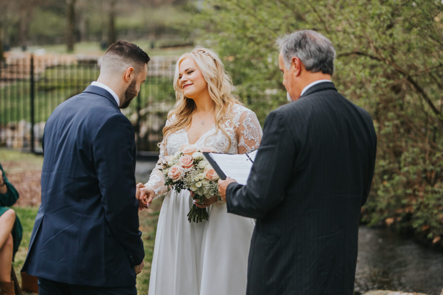 Elopement Wedding A couple exchanges vows outdoors, with the bride looking at the groom while holding a bouquet of flowers. The bride is in a white dress and the groom in a navy blue suit. An officiant, dressed in a black suit and holding a script, stands beside them. Greenery and a fence are visible in the background, capturing the essence of an intimate elopement. Elopements Inc