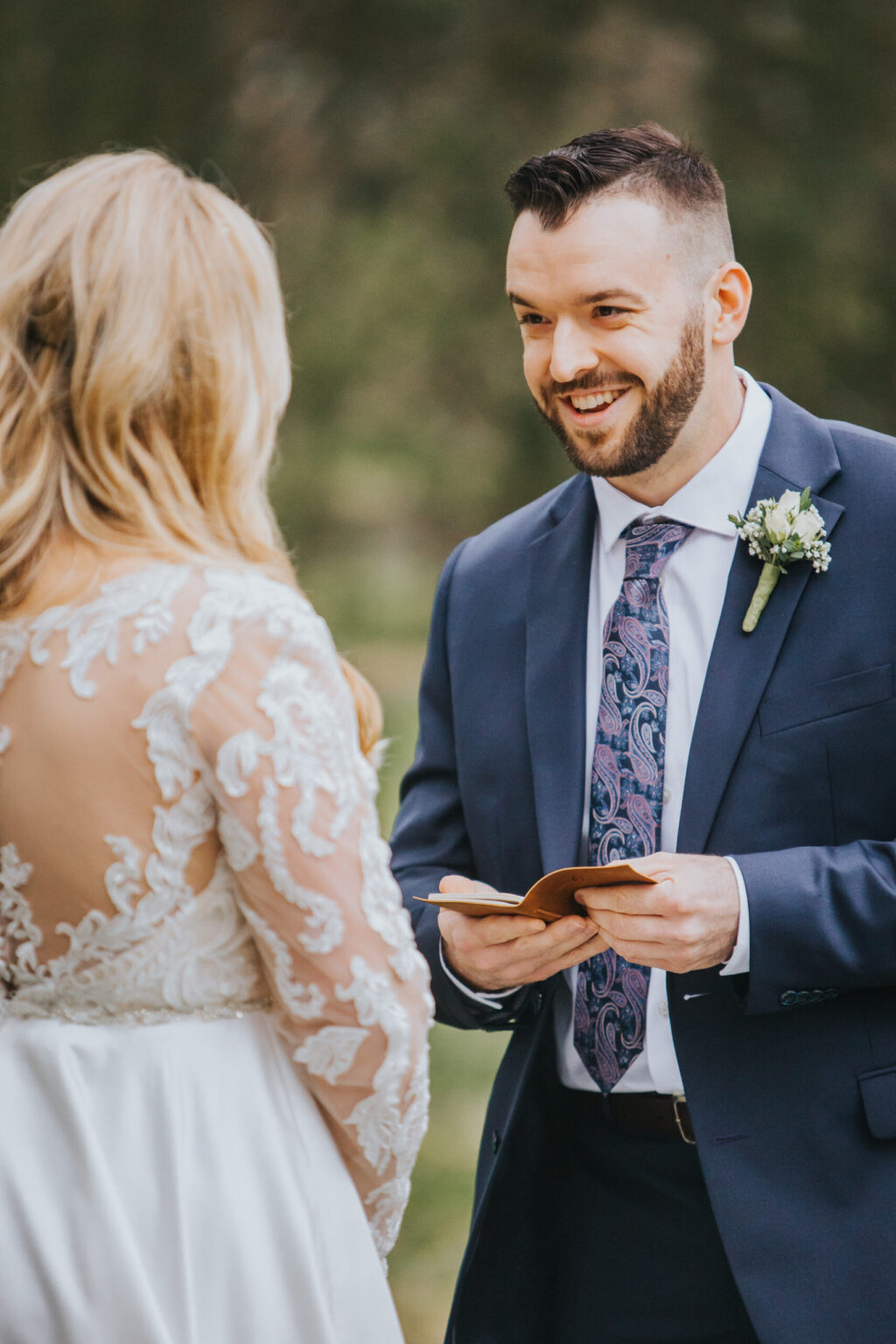 Elopement Wedding A groom in a navy suit and patterned tie reads vows from a small book to his bride, seen from the back in a long-sleeve, sheer white lace wedding dress. They stand outdoors during their intimate elopement, greenery blurred in the background as they smile at each other. Elopements Inc