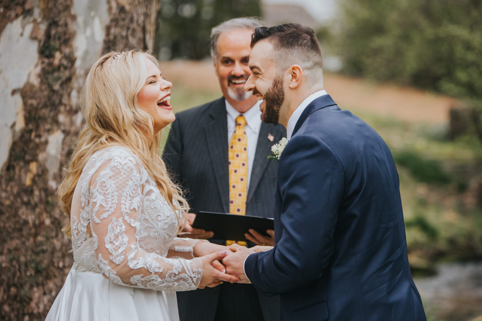 Elopement Wedding A couple laughs joyfully while holding hands during their intimate elopement ceremony. The bride wears a long-sleeved lace gown, and the groom sports a navy suit with a white shirt. An officiant in a dark suit stands behind them, holding a book. Trees and greenery are visible in the background. Elopements Inc