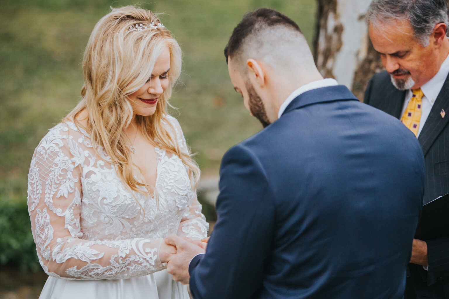 Elopement Wedding A bride in a white lace gown and jeweled headpiece holds hands with a groom in a navy blue suit while an officiant in a suit and yellow tie conducts their intimate elopement ceremony outdoors. The bride smiles and looks down at their hands. Trees and greenery are visible in the background. Elopements Inc