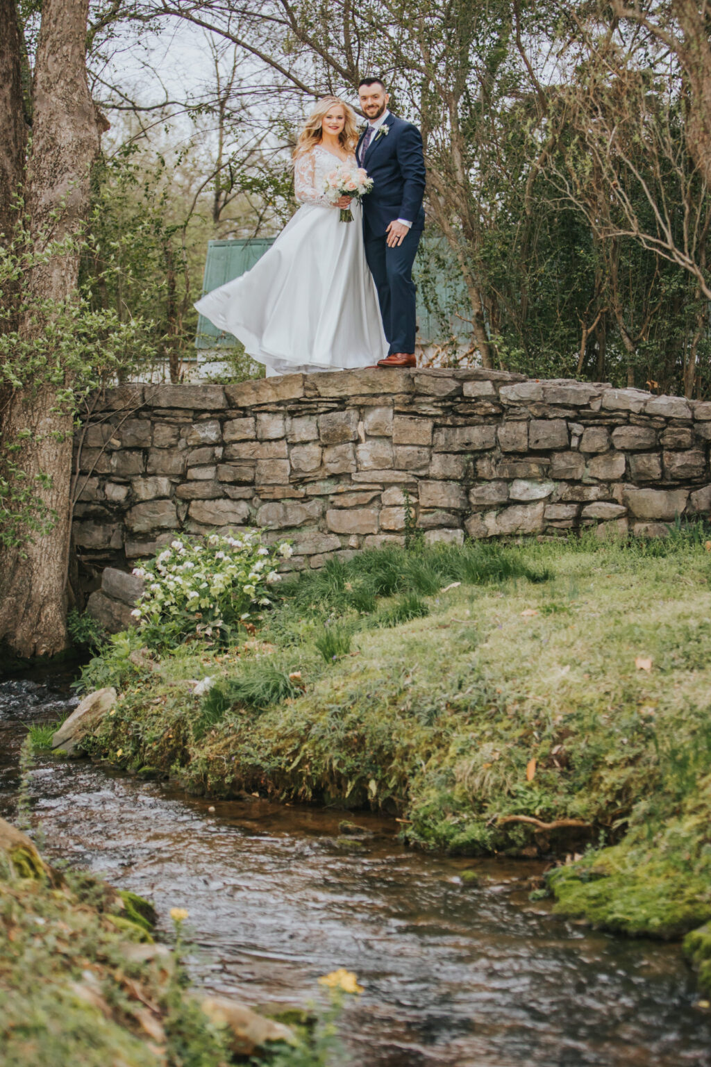 Elopement Wedding A couple stands on a stone bridge above a small creek, surrounded by greenery. The woman wears a white wedding dress and holds a bouquet, while the man is in a dark suit with a tie. Both smile, looking happy, as their elopement unfolds. Trees and shrubs frame the scene, creating a vibrant and picturesque setting. Elopements Inc