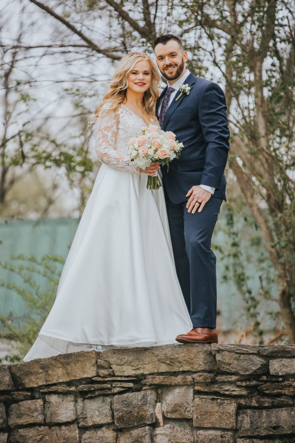 Elopement Wedding A bride and groom stand outdoors on a stone ledge, posing for a photo. The bride, in a long white dress with lace details and holding a bouquet of light-colored flowers, gazes lovingly at her groom. Dressed in a navy suit with a light blue tie and boutonniere, they epitomize the simplicity of an elopement. Trees and a blurred fence are in the Elopements Inc