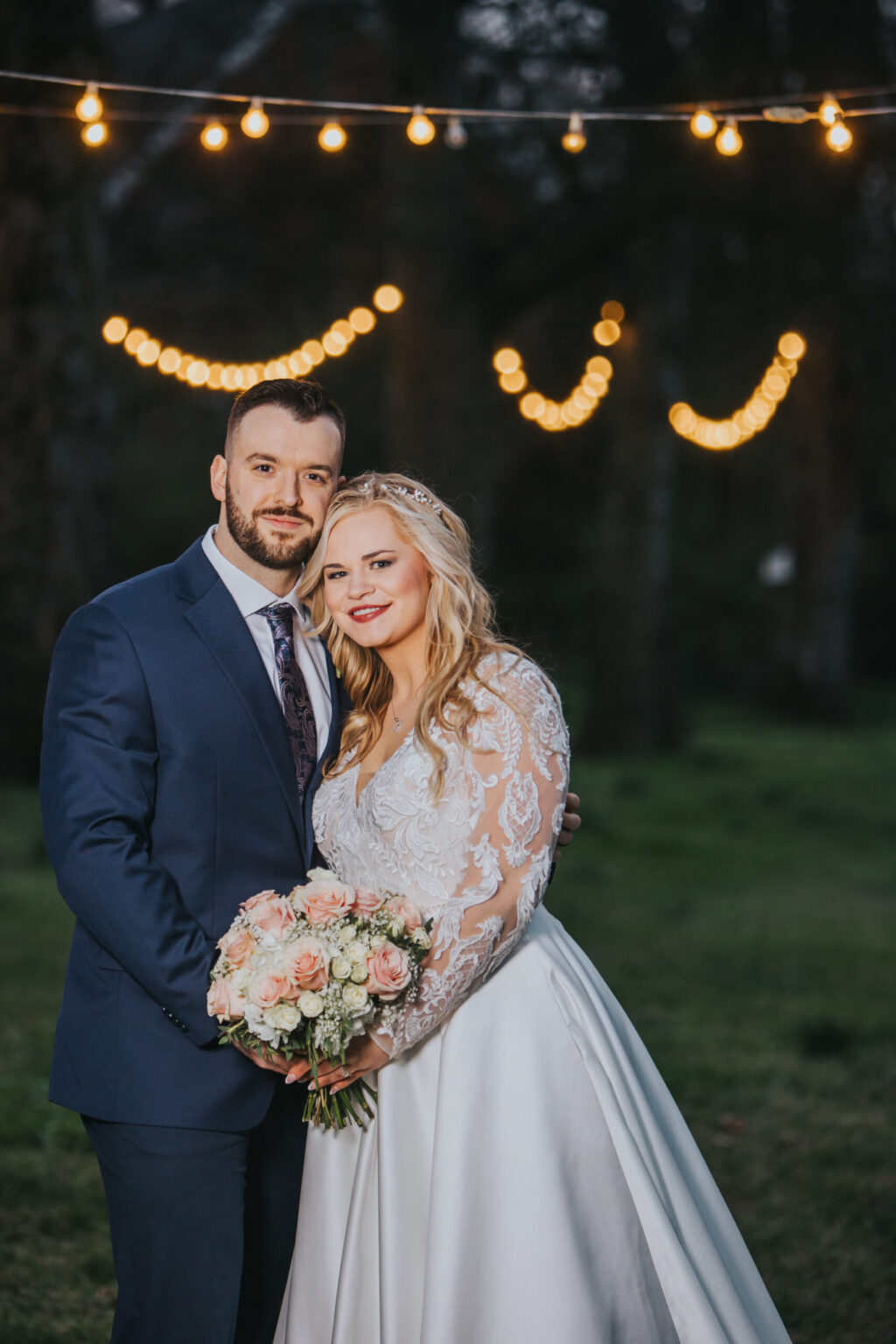 Elopement Wedding A bride and groom pose outdoors under string lights at dusk, looking as if they’ve just eloped. The bride, in a long lace-sleeved white gown, holds a bouquet of pastel flowers and smiles. The groom, dressed in a navy suit with a white shirt and dark tie, stands beside her with his arm around her waist amid lush greenery. Elopements Inc