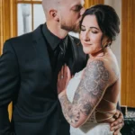 Elopement Wedding A groom in a black suit and tie kisses the forehead of a bride in a sleeveless white lace dress. The bride, with extensive floral tattoos on her exposed arm, has her eyes closed and a slight smile. They are standing closely in an indoor setting with wooden accents and large windows, celebrating their intimate elopement. Elopements Inc