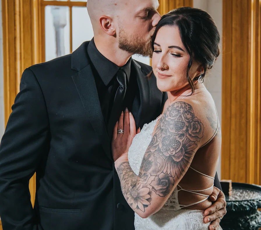 Elopement Wedding A groom in a black suit and tie kisses the forehead of a bride in a sleeveless white lace dress. The bride, with extensive floral tattoos on her exposed arm, has her eyes closed and a slight smile. They are standing closely in an indoor setting with wooden accents and large windows, celebrating their intimate elopement. Elopements Inc