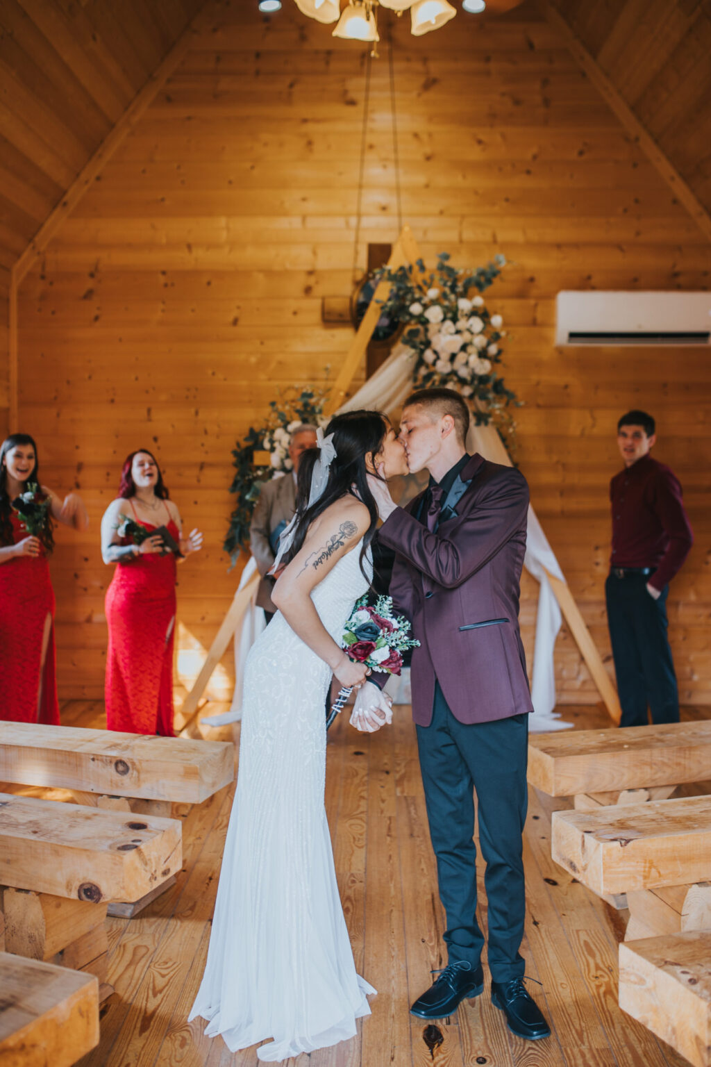 Elopement Wedding A bride in a white dress and a groom in a maroon suit kiss at the altar in a wooden chapel. Embracing the spirit of elopements, the bride holds a bouquet while the groom places his hand on her waist. Bridesmaids in red dresses and groomsmen in dark suits stand behind them, clapping and smiling in celebration. Elopements Inc