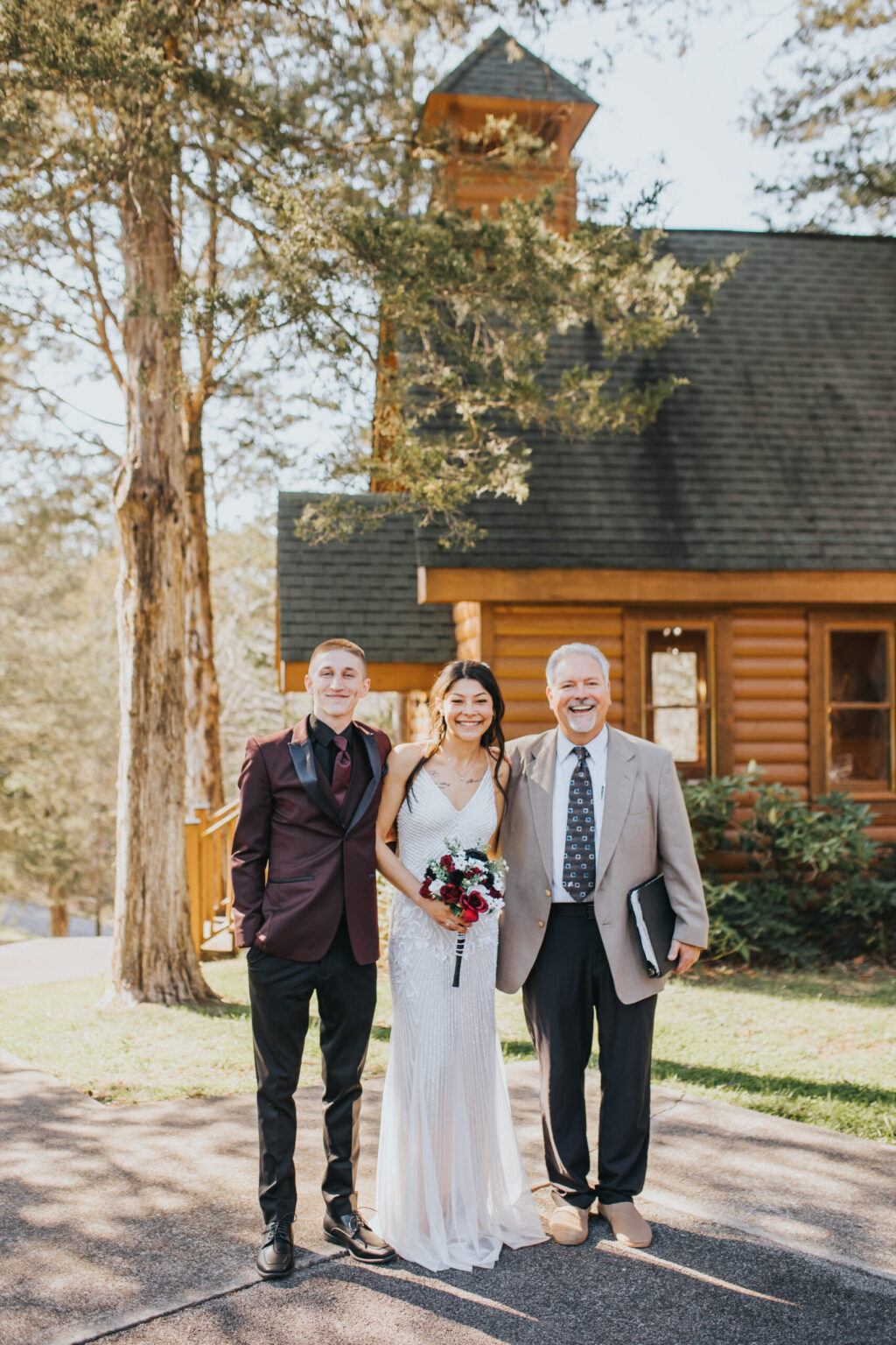 Elopement Wedding Three people stand outside in front of a log cabin surrounded by trees. A bride in a white gown holds a bouquet of red and white flowers, flanked by two men – one in a dark suit and burgundy jacket, and the other in a beige suit with a book and folder, capturing the essence of an intimate elopement. Elopements Inc