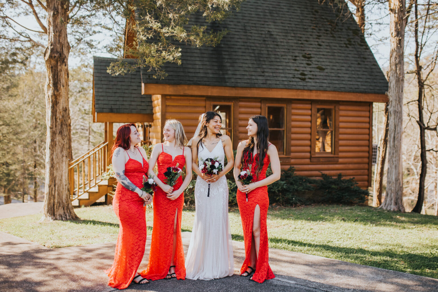 Elopement Wedding Four women stand in front of a wooden cabin in a wooded area. One woman, in a white dress, holds a bouquet and stands between three women in matching red dresses who also hold bouquets. They all smile and appear to be in a celebratory mood under the sunlight, as if they've just eloped to this serene spot. Elopements Inc