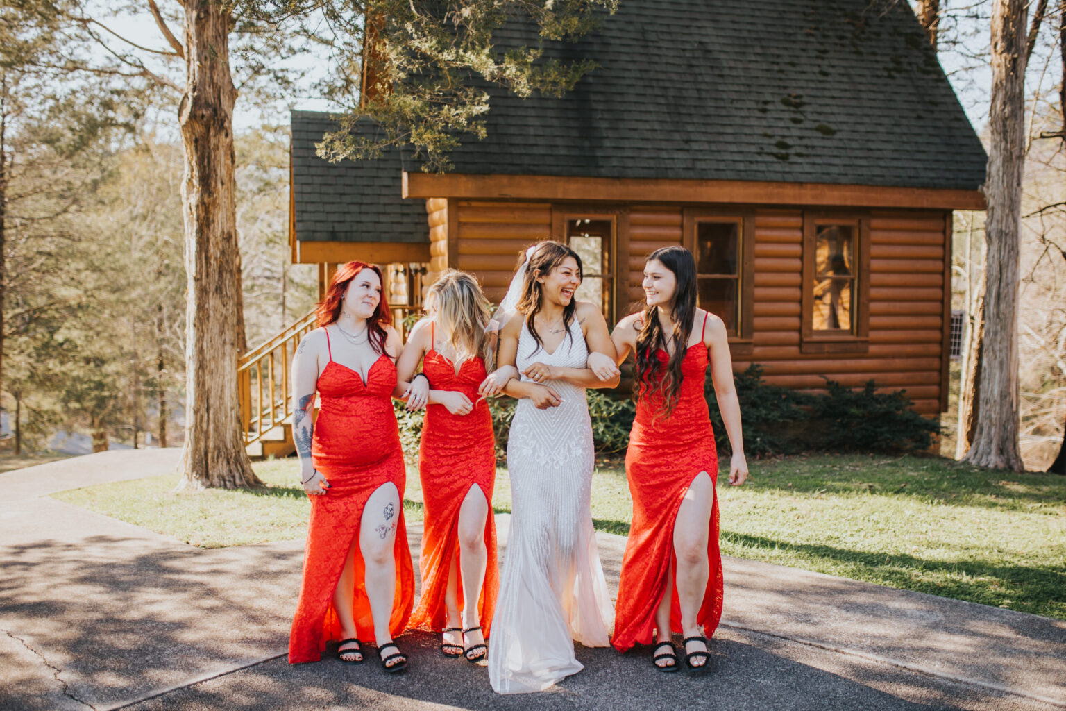 Elopement Wedding Four women walk arm-in-arm on a paved path in front of a wooden cabin. Three wear red dresses with thigh-high slits, black shoes, and have either red or blonde hair, while the fourth, in the center, wears a white dress with a lace overlay and has brown hair. Trees surround the cabin and path, perfect for an intimate elopement. Elopements Inc