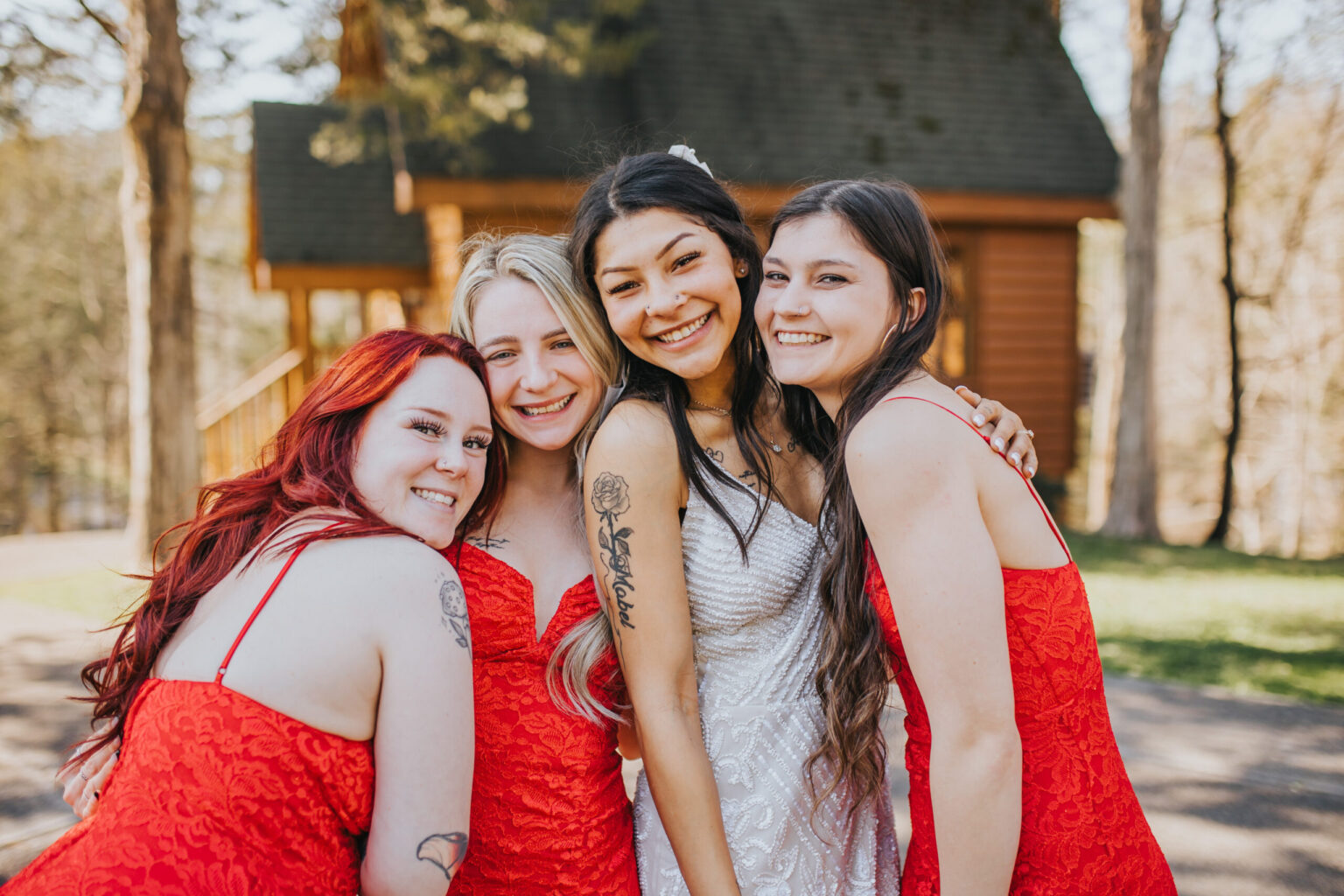 Elopement Wedding Four women are posing closely together outdoors, smiling at the camera. The woman in the center, wearing a white dress, appears to be a bride eloping. The other three women in matching red dresses proudly display tattoos on their arms. Trees and a wooden building are visible in the background. Elopements Inc