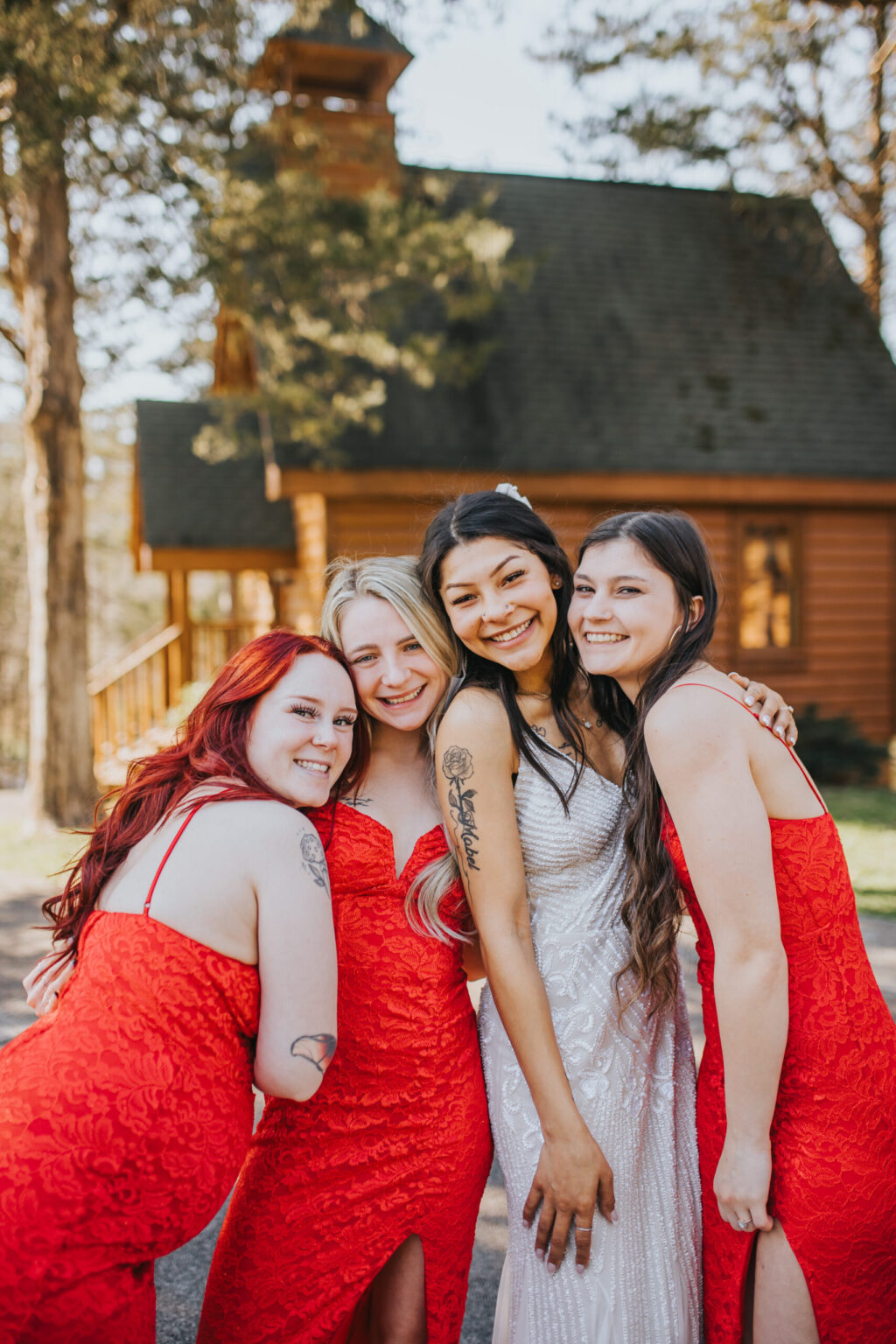 Elopement Wedding Four smiling women pose together outdoors, in front of a wooden cabin surrounded by trees. One, wearing a white wedding dress, is flanked by three others in red lace dresses with side slits. All have long hair; the bride and two bridesmaids have it down, a third wears a ponytail. This intimate elopement radiates joy and unity. Elopements Inc