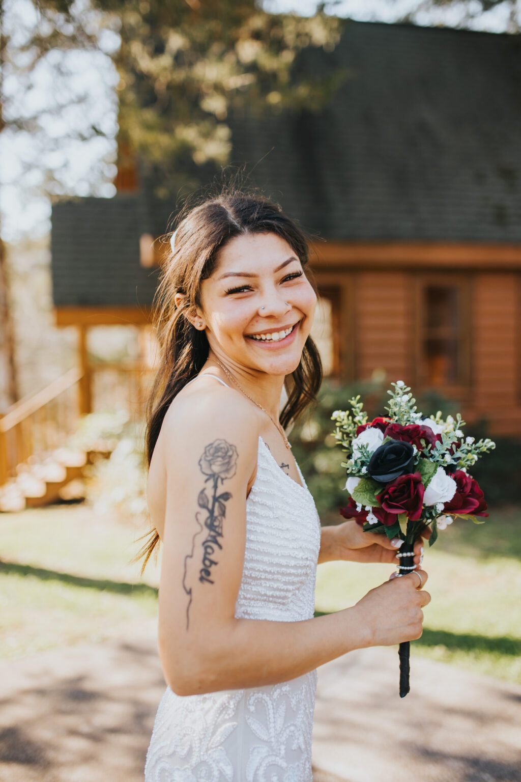 Elopement Wedding A bride with long dark hair and a tattoo on her upper right arm holds a bouquet of red, white, and black flowers. She is wearing a white sleeveless lace wedding dress and smiling broadly. The backdrop reveals a wooden cabin and sunlit trees, perfect for their intimate elopement. Elopements Inc