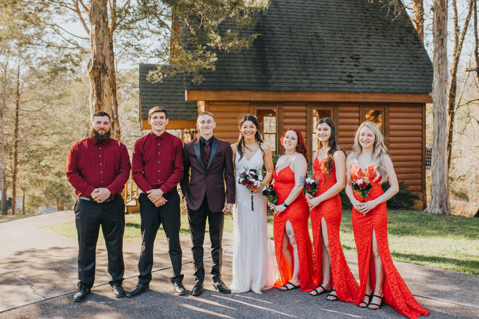 Elopement Wedding A group of people posed in front of a log cabin. Three men stand on the left, dressed in formal attire, two in red shirts and one in a dark suit. Three women stand on the right, wearing red dresses and holding bouquets. Trees and grass surround the cabin in the background, creating an intimate elopement setting. Elopements Inc